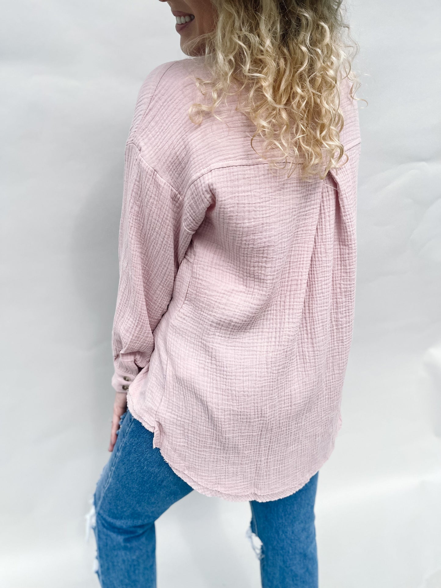 Back view of a fashionable model wearing a light pink linen button-up shirt with a longer back that covers the bottom, paired with jeans and a trendy hat. The model looks back over her shoulder with a happy smile