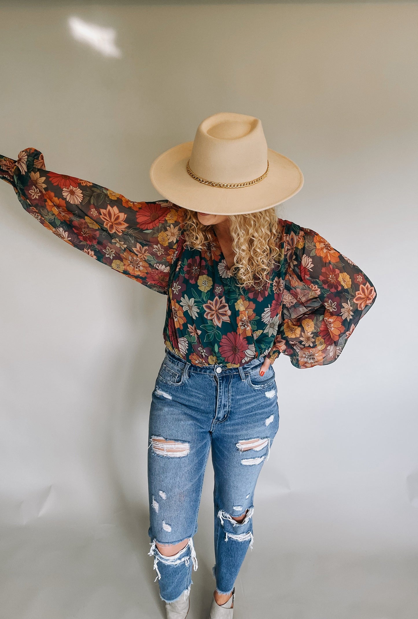 Stylish model wearing a vibrant green, orange, and red floral bodysuit with flowy sleeves, paired with blue jeans and a wide-brimmed hat with a gold chain accent