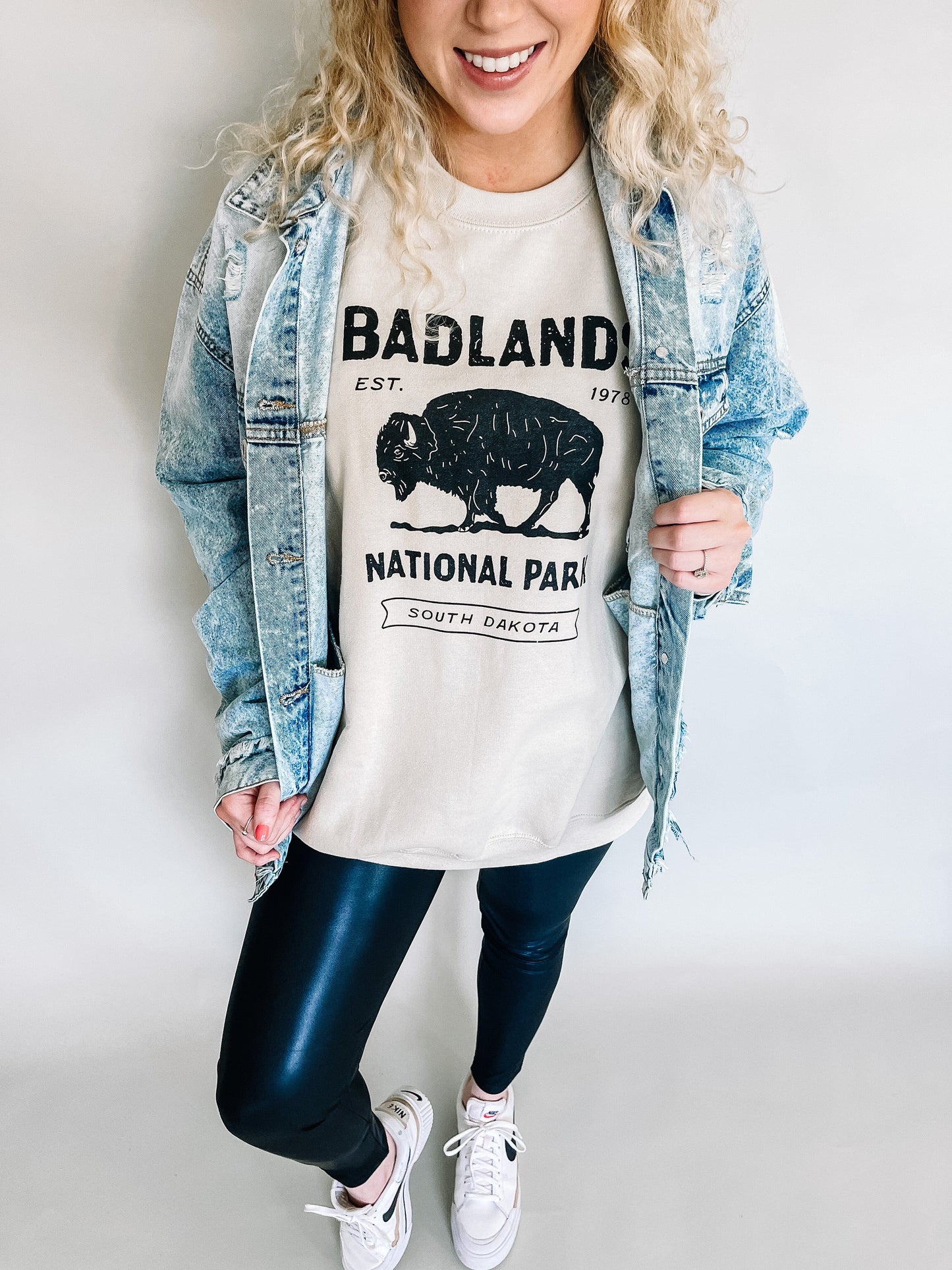 Casual yet stylish model wearing an oversized taupe sweatshirt with a graphic of Badlands National Park and a bison, paired with sleek leather leggings and comfortable white Nike tennis shoes, creating a trendy and relaxed outfit