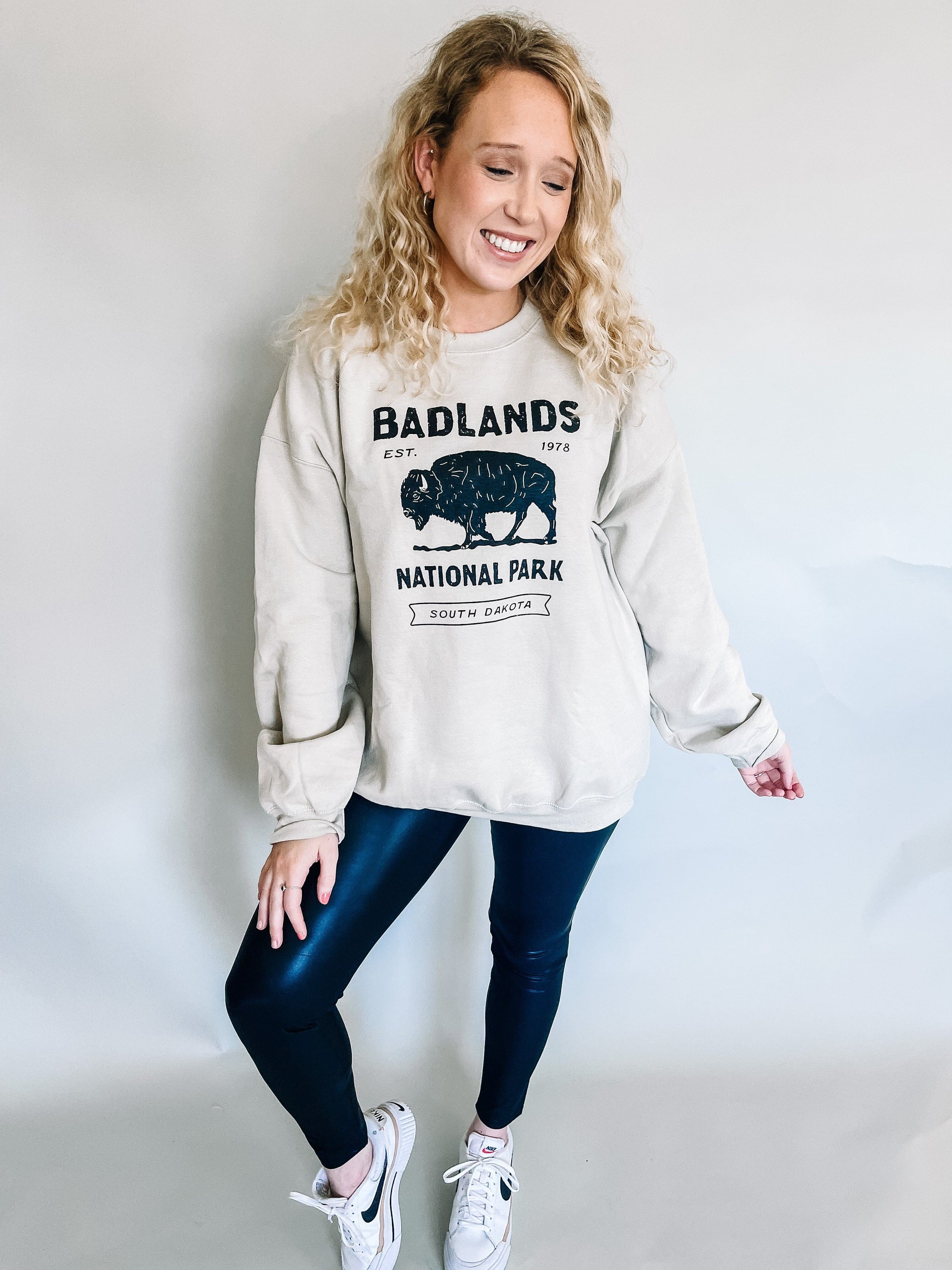 Casual yet stylish model wearing an oversized taupe sweatshirt with a graphic of Badlands National Park and a bison, paired with sleek leather leggings and comfortable white Nike tennis shoes, creating a trendy and relaxed outfit